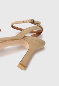 Beige suède hoge hak met een smalle, gebogen hak en een tan enkelbandje met een metalen gesp. Gladde textuur, minimalistisch ontwerp.
