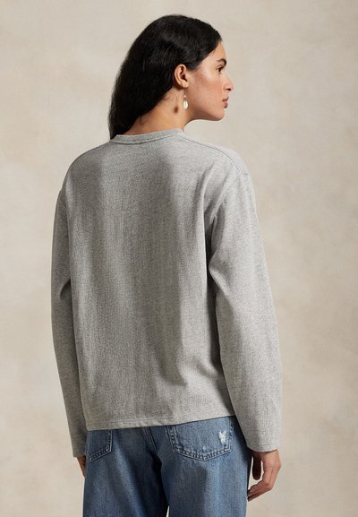 Donna con lunghi capelli scuri che indossa una maglia grigia a maniche lunghe e jeans blu, vista da dietro su uno sfondo neutro.