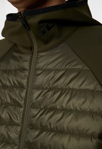 Framifrån vy av en grön vadderad jacka med dragkedja och uppfällt krage som delvis täcker nacken, bärs av en person.