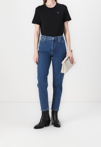 Černé bavlněné tričko, modré džíny s vysokým pasem, kotníkové černé boty na podpatku, držící béžovou kabelku. Jednoduchý design, bez vzorů.