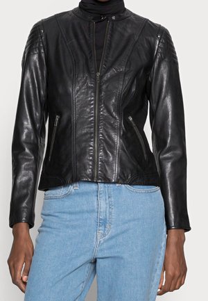 Personne portant une veste en cuir noire ajustée avec des poches zippées et un jean bleu taille haute sur un fond uni.