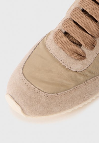 Närbild av en beige sneaker-tå med mocka och tygmaterial samt flätade beige snören mot en enkel bakgrund.