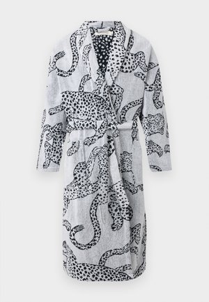 Άνδρας στέκεται ξυπόλητος φορώντας ένα λευκό μπουρνούζι μέσου μήκους με μαύρο αφηρημένο λεοπάρ σχέδιο και έχει τα χέρια στις μπροστινές τσέπες.