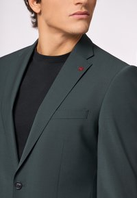 Blazer verde scuro con un design strutturato, chiusura a un bottone e un accento ricamato rosso sul rever, indossato sopra una camicia nera.
