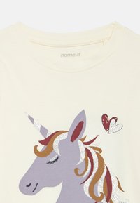 Maglietta color crema chiaro con grafica di un unicorno viola, corno e criniera scintillanti in argento, con tonalità di marrone, bianco e rosso. Due cuori sopra.