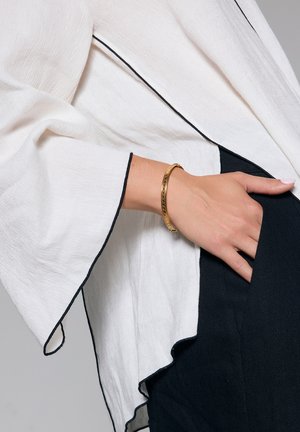 Biała bluzka z czarnymi wykończeniami, z szerokimi rękawami. Złota bransoletka zdobi rękę spoczywającą w kieszeni z czarnego materiału.