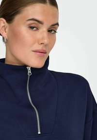 Pull taille zippée en tissu bleu marine avec col haut, dotée d'une fermeture éclair argentée et de détails de couture minimalistes. Texture lisse et design décontracté.