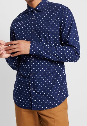 Hombre vestido con camisa azul marino con botones y un patrón pequeño de cuadros blancos y pantalones marrones sobre un fondo liso.