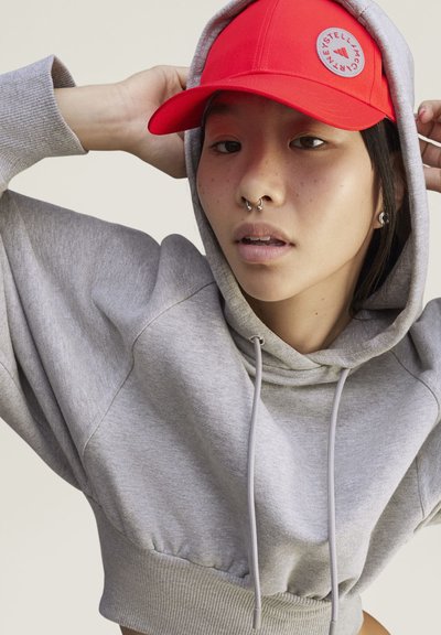 Joven con un pendiente en la nariz que lleva una sudadera gris con capucha y una gorra roja brillante con un logo circular ajustándose la gorra.