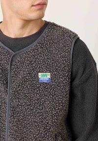 Siva, teksturirana flis jopica z okrolim izrezom na vratu, črnim gumbom za zapiranje in pisano značko z logotipom na prsih.