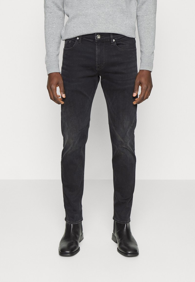 Jeans neri slim-fit con una texture liscia, dotati di cinque tasche, chiusura frontale con bottone e dettagli di leggera scoloritura. Indossati con stivali neri.