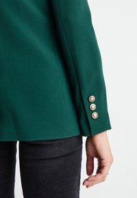 Zelený blazer s texturovanou látkou. Má přizpůsobený střih a tři akcenty zlatých knoflíků na manžetě. V kompletu s tmavými džíny.