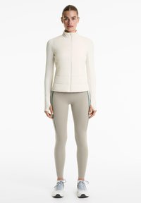 Gilet matelassé crème avec col haut, associé à des leggings beiges avec des rayures latérales teal, et des chaussures de sport bleu clair.