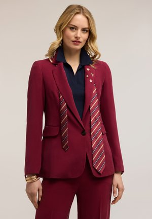 Donna che indossa un completo bordeaux con cravatte a righe che pendono dal blazer, camicia blu navy e braccialetti dorati, in piedi contro uno sfondo neutro.
