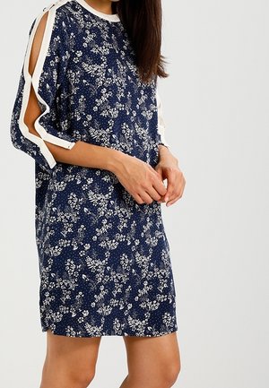 Femme portant une robe bleu marine à fleurs avec des manches épaules dénudées bordées de blanc et nouées avec des nœuds, debout devant un fond clair uni.