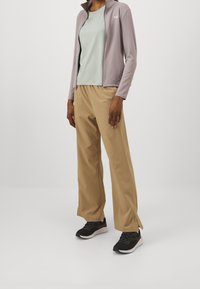 Ljusgrå zip-up jacka över en blekgrön t-shirt, kombinerad med beige vida byxor och svarta sportskor, med en slank design.