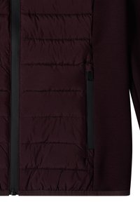 Gros plan d'une veste bordeaux foncé montrant un panneau avant matelassé avec une fermeture éclair noire verticale et une manche en tissu lisse.
