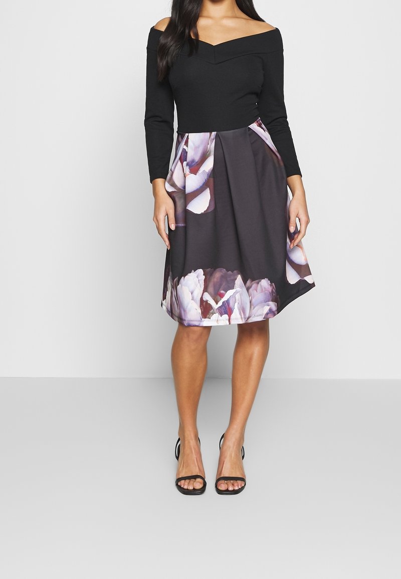 Femme portant un haut noir épaules dénudées et une jupe noire longueur genoux avec un grand motif floral violet clair, et des talons ouverts noirs.