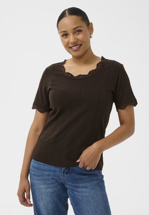 Femme aux cheveux bruns portant un haut marron à manches courtes avec un bord en dentelle et un jean bleu, souriant, une main reposant près de sa taille.