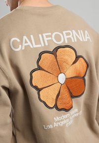 Beige sweatshirt featuring “CALIFORNIA” in white text. Back displays a large orange flower design with brown outlines and white center detail.