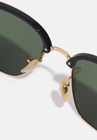 Sunglasses with black acetate frames, green lenses, and gold metal accents. Features include a bridge with engraved details and beige nose pads.
