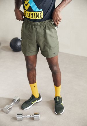 Personne en short de sport ajustant la taille, portant des chaussettes jaunes et des baskets vertes Nike, debout près d’haltères et d’un ballon médicinal au sol.