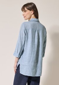 Camicia a righe blu e bianche con colletto, maniche lunghe arrotolate fino al gomito e una plissettatura sul retro, realizzata in tessuto leggero.