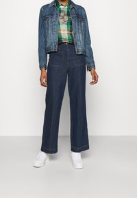 Chaqueta de mezclilla con un lavado azul claro, combinada con una camisa de cuadros de botones y jeans de pierna ancha oscuros, rematada con zapatillas blancas.