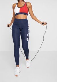 Biustonosz sportowy z czerwonymi, białymi i granatowymi blokami kolorów; granatowe legginsy z wysokim stanem z napisem "TOMMY SPORT"; osoba trzymająca skakankę.