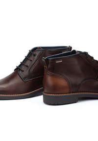 Botas de tobillo de cuero marrón con una textura suave, que presentan un diseño de cordones, acentos en un tono marrón claro y una suela de goma resistente.