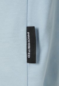 Una tela de color azul claro con una textura suave presenta una etiqueta negra vertical cosida en el lateral, que muestra la palabra "PROHIBIDO" en blanco.