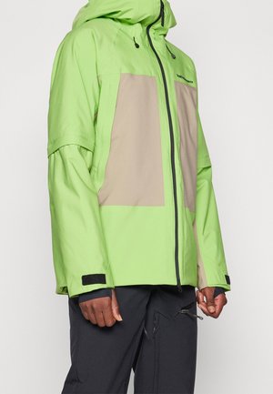 Personne portant une veste à capuche vert citron et beige avec une fermeture éclair noire et un pantalon noir avec des poches zippées.
