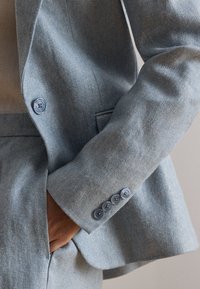 Käsi helehallis tekstuuriga ülikonnas, mille varrukal on neli nööpi ja käsi puhkab ülikonnataskus.