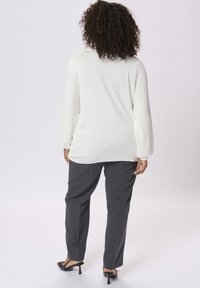 Frau mit lockigem Haar, die einen weißen Pullover mit langen Ärmeln, graue Hosen und schwarze Absatzschuhe trägt, steht von der Kamera abgewandt vor einem schlichten Hintergrund.
