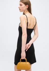 Νεαρή γυναίκα με μαύρο αμάνικο φόρεμα κρατά μικρό καφέ τσαντάκι πίσω από την πλάτη της, φορά ένα μενταγιόν σκουλαρίκι.