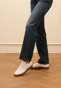 White leather sneakers with a textured finish, featuring a rubber gum sole and subtle side pattern, worn with dark denim jeans.