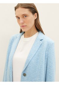 Light blue textured blazer with notched lapels and a single button closure, worn over a white top. Focus on tailored fit and fabric detail.