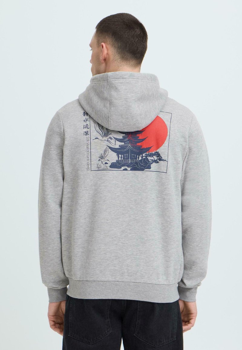 Homme portant un sweat à capuche gris avec un graphisme de pagode japonaise, de grues et de soleil rouge dans le dos, debout devant un fond clair uni.