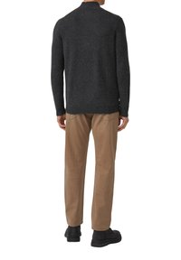 Uomo che indossa un maglione grigio scuro, pantaloni beige e scarpe nere, in piedi rivolto di spalle su sfondo bianco.