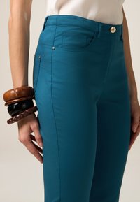 Teal broeken van gladde stof met een aansluitend ontwerp, voorzien van voorzakken en een decoratieve gouden knop in de taille.