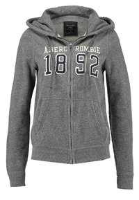 Sudadera gris con cremallera, hecha de un tejido suave. Presenta un bolsillo frontal, capucha con cordón y estampado "Abercrombie 1892" en el pecho.