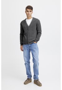 Cardigan gris de punto con escote en V y botones en la parte delantera, llevado sobre una camiseta blanca. Combinado con jeans de mezclilla azul claro y zapatos de ante marrones.