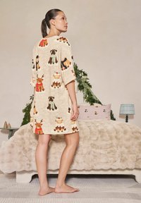 Žena v béžových šatách s dovolenkovými motívmi a zvieracími potlačami stojí naboso vedľa postele s huňatou prikrývkou a sviatočnou zeleňou.