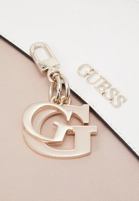 Matière en cuir rose ornée d'un pendentif logo "G" en métal doré avec des anneaux métalliques reliés. Une section blanche affiche le nom "GUESS" en lettres dorées.