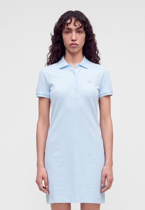 Femme aux longs cheveux bouclés portant une robe polo bleu clair à manches courtes, debout devant un fond uni.