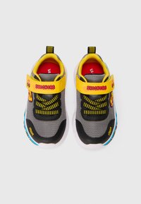 Sapatos atléticos cinzentos com um cabedal em malha respirável, tira de Velcro amarela, detalhes pretos e uma sola colorida.