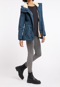 Chaqueta de invierno verde azulado con forro de piel sintética, cierres de cremallera y a presión, capucha ajustable, combinada con jeans ajustados grises y botas negras de tobillo.