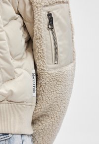 Párnázott bézs dzseki bolyhos textúrájú anyaggal az ujjakon, cipzáras zseb és bordás mandzsetta található rajta. A logócímke a oldalvarrásnál látható.