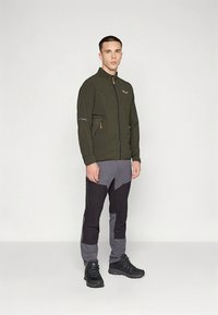 Lett olivengrønn jakke med glidelås, grå bukser med svarte detaljer, og svarte tursko, stående foran en hvit bakgrunn.