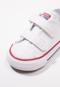 Valge nahkne tennis, millel on kaks Velcro-rihma, punase kummi allaotuse aktsent, ümar toe-kapp ja tekstuuriga külgedetailid. Klassikaline disain.